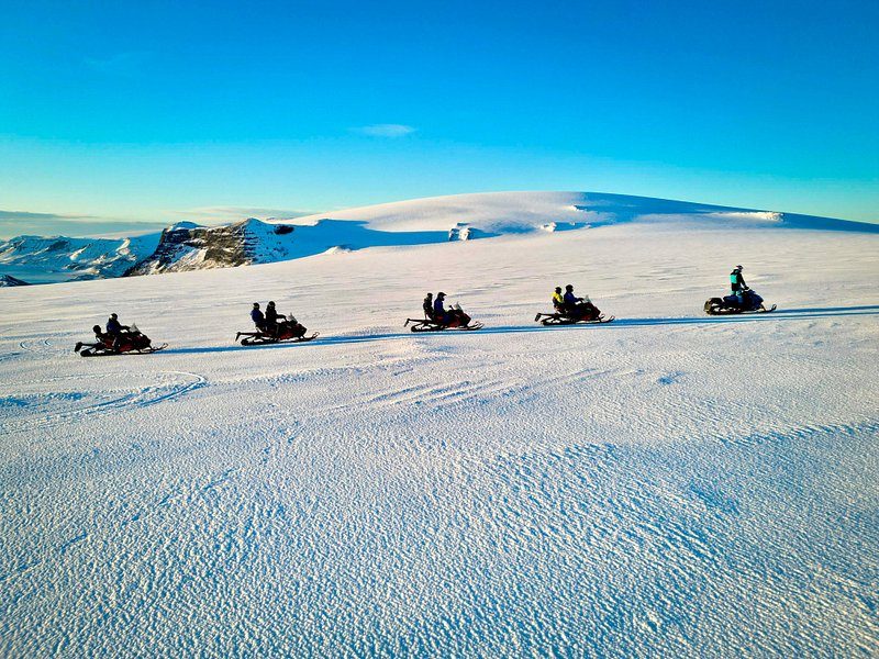 A line of five snowmobiles crossing a flat white ice cap under a blue sky