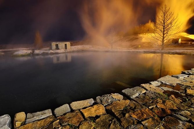 A steaming geothermal pool at night beside a small stone hut and a lit tree