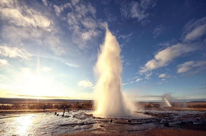 Strokkur erupting against a low sun, visitors small at the edge of the field
